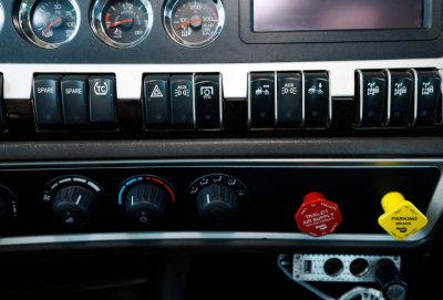 Cockpit dashboard of a Kenworth truck showing gauges, switches, and control knobs with circular gauges and warning/toggle buttons.