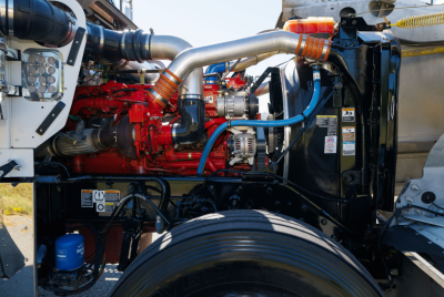 Kenworth W900L engine bay showing red diesel engine, silver intake pipes, blue hoses, black radiator, and yellow-orange clamps