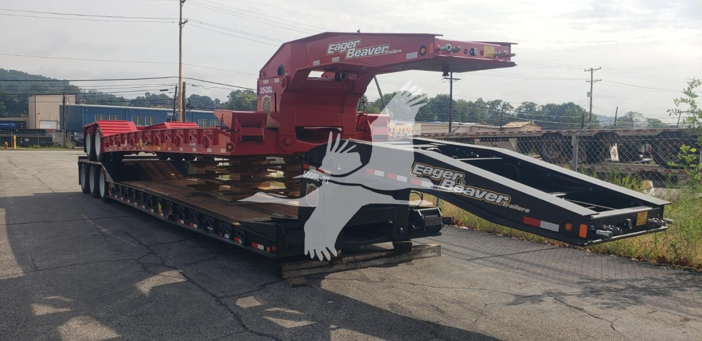Trailer in a transport yard showing a red gooseneck lowboy trailer with foldable ramps and a black loading ramp at the back.
