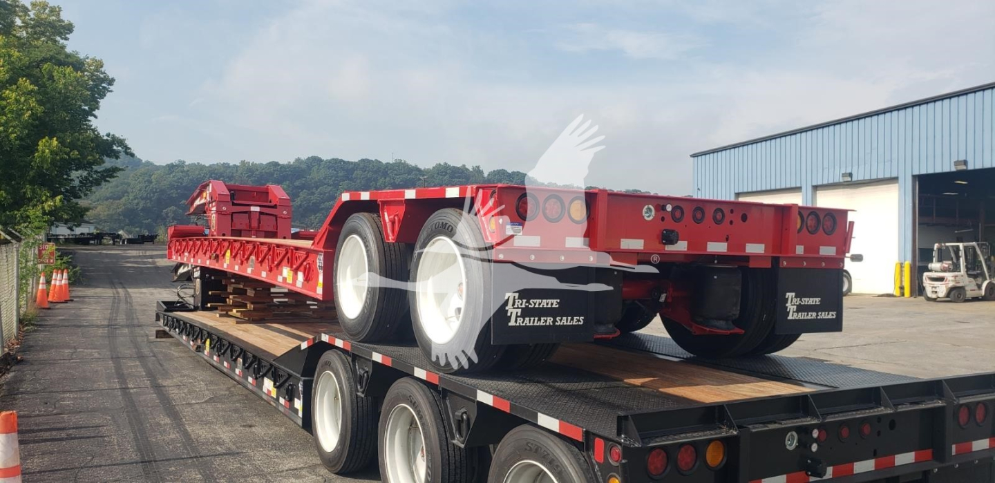 Trailer on a flatbed trailer showing a red gooseneck lowboy with dual rear axles and large tires. Visible text includes VL088753 stock tag and brand logos on the rear.