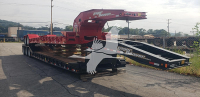 Trailer in a transport yard showing a red gooseneck lowboy trailer with foldable ramps and a black loading ramp at the back.