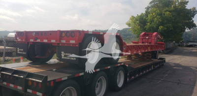 Trailer showing a red lowboy flatbed with wheel wells and a detachable third axle section, branding and VIN tag on the side; overall scene outdoors.