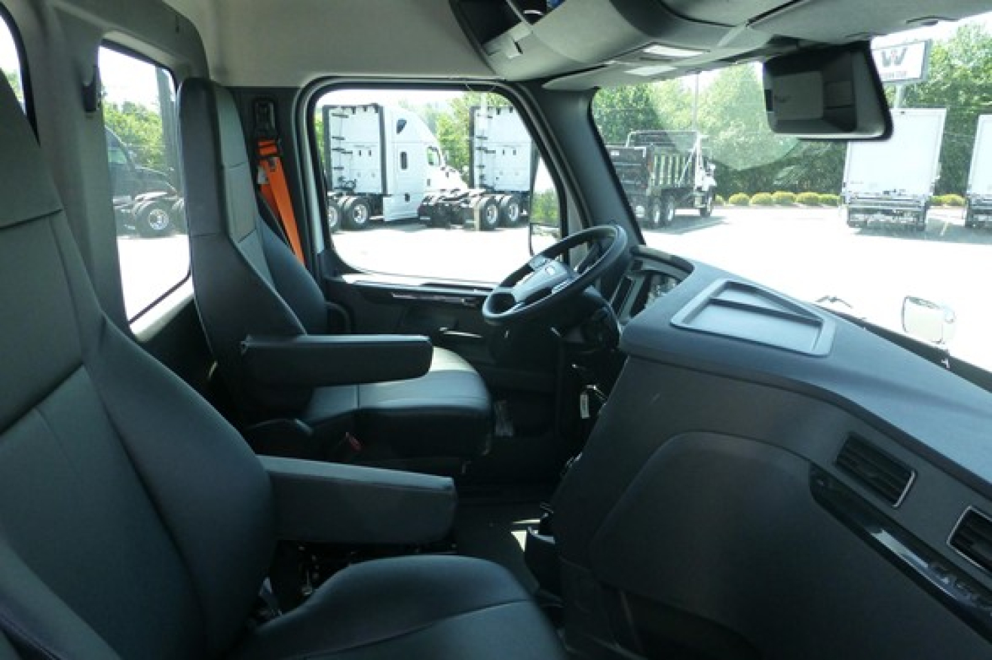 Truck cab interior showing steering wheel, dashboard, and driver seat with armrest; light gray/black plastics; windshield and side window visible.