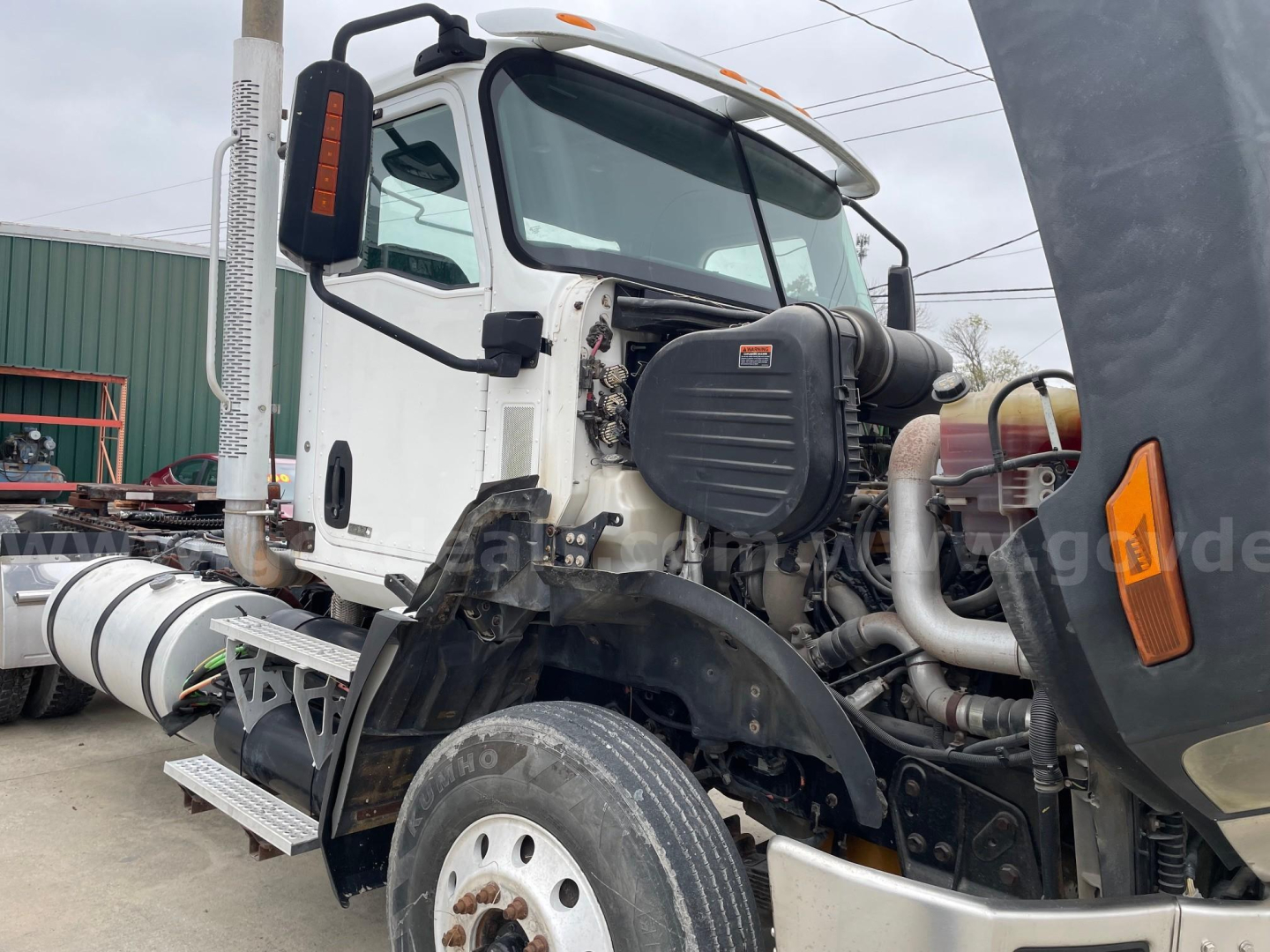 Front-left view of a white Caterpillar truck cab with exposed engine components and large exhaust stack on the side; visible tires, part of the chassis, and engine bay area.