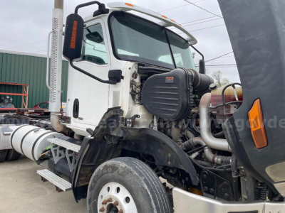 Front-left view of a white Caterpillar truck cab with exposed engine components and large exhaust stack on the side; visible tires, part of the chassis, and engine bay area.
