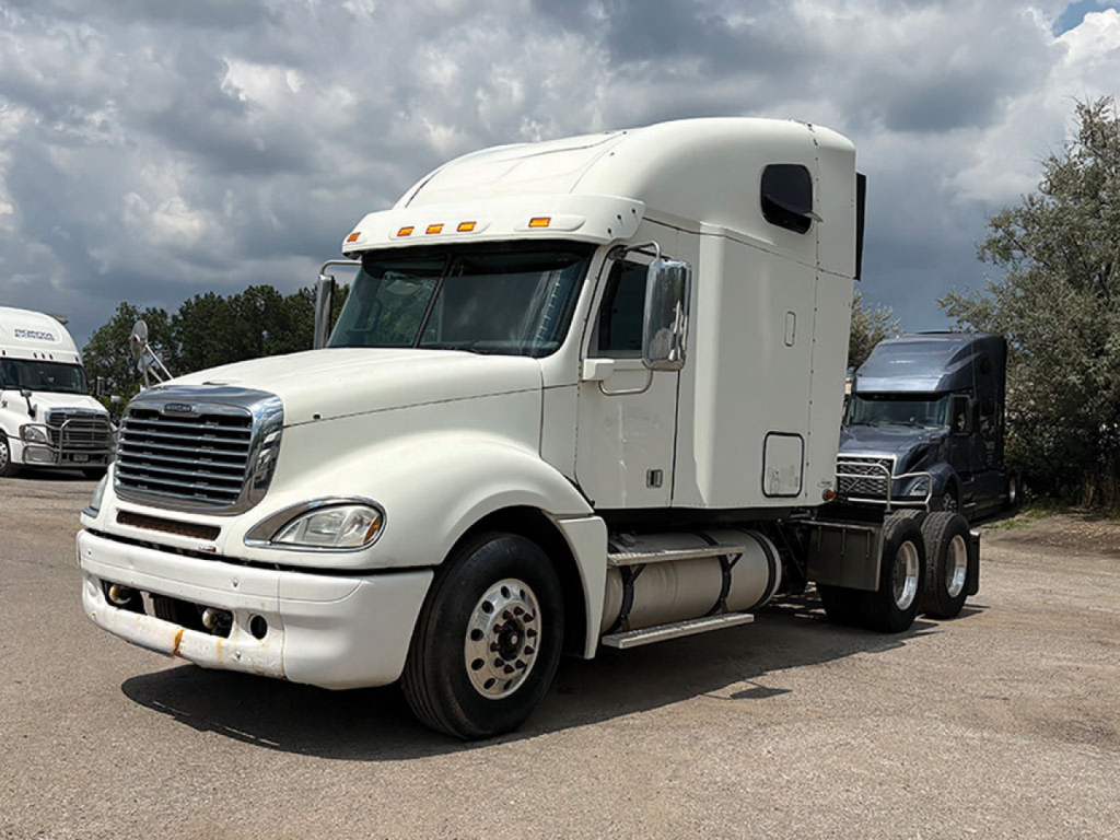 White Freightliner CL120 Columbia sleeper cab truck in outdoor lot. Front bumper and grille visible, fuel tanks under cab, large side mirrors, aerodynamic sleeper curvature.
