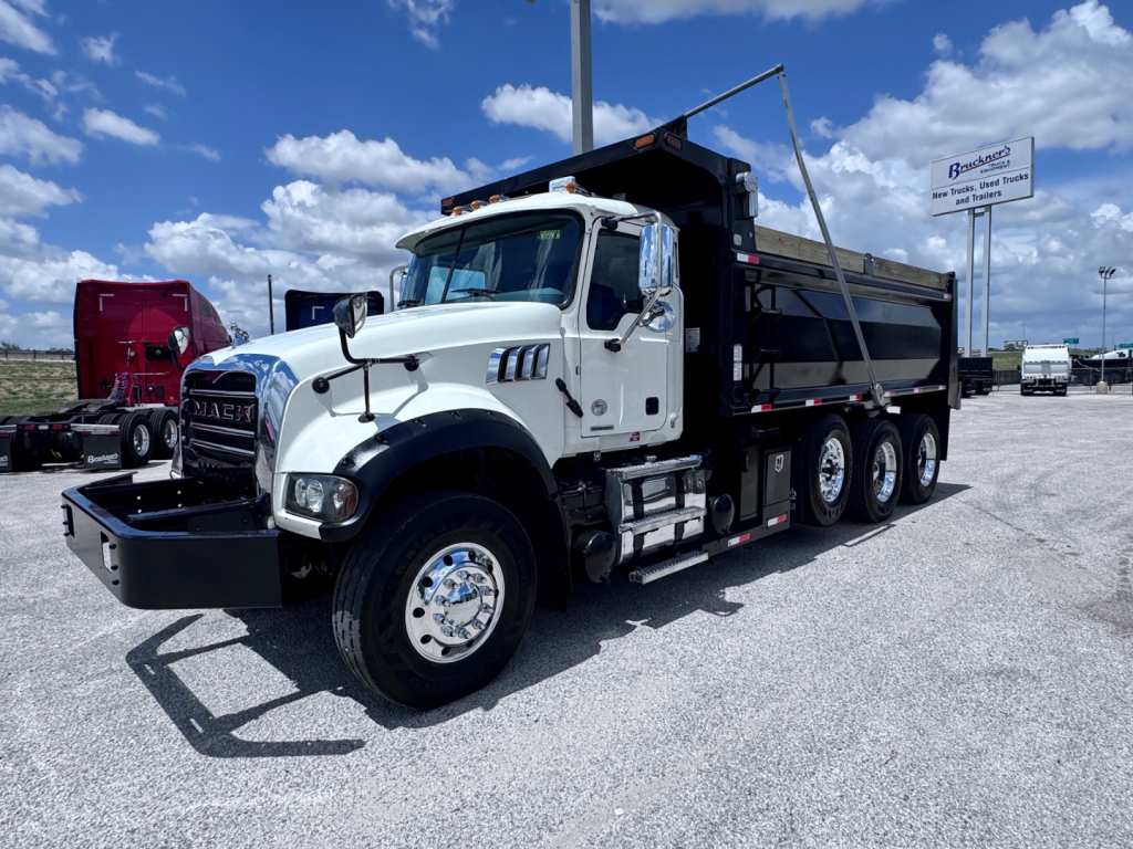 Truck cab and dump truck bed of a 2023 Mack GR84F in-service showing front grille, dual rear axles, white cab, and black dump body with side railing.