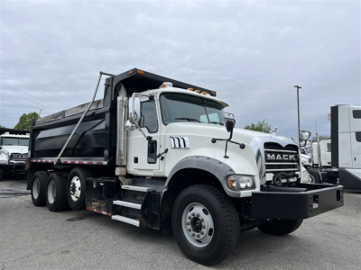 Dump truck showing 2023 Mack GR84F with white cab and black dump bed, side steps, grille, and front bumper; visible cab details and dual rear axles.