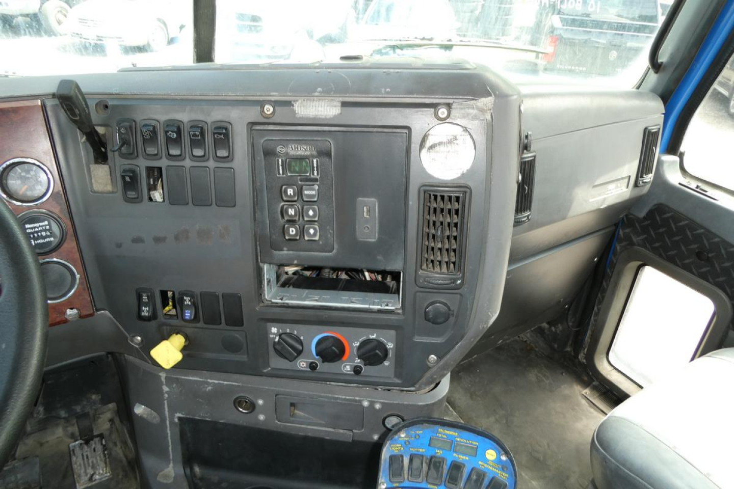 Dashboard and center console of a Mack mixer truck cab showing control panels, switches, gauges, and a circular blue device on the floor.