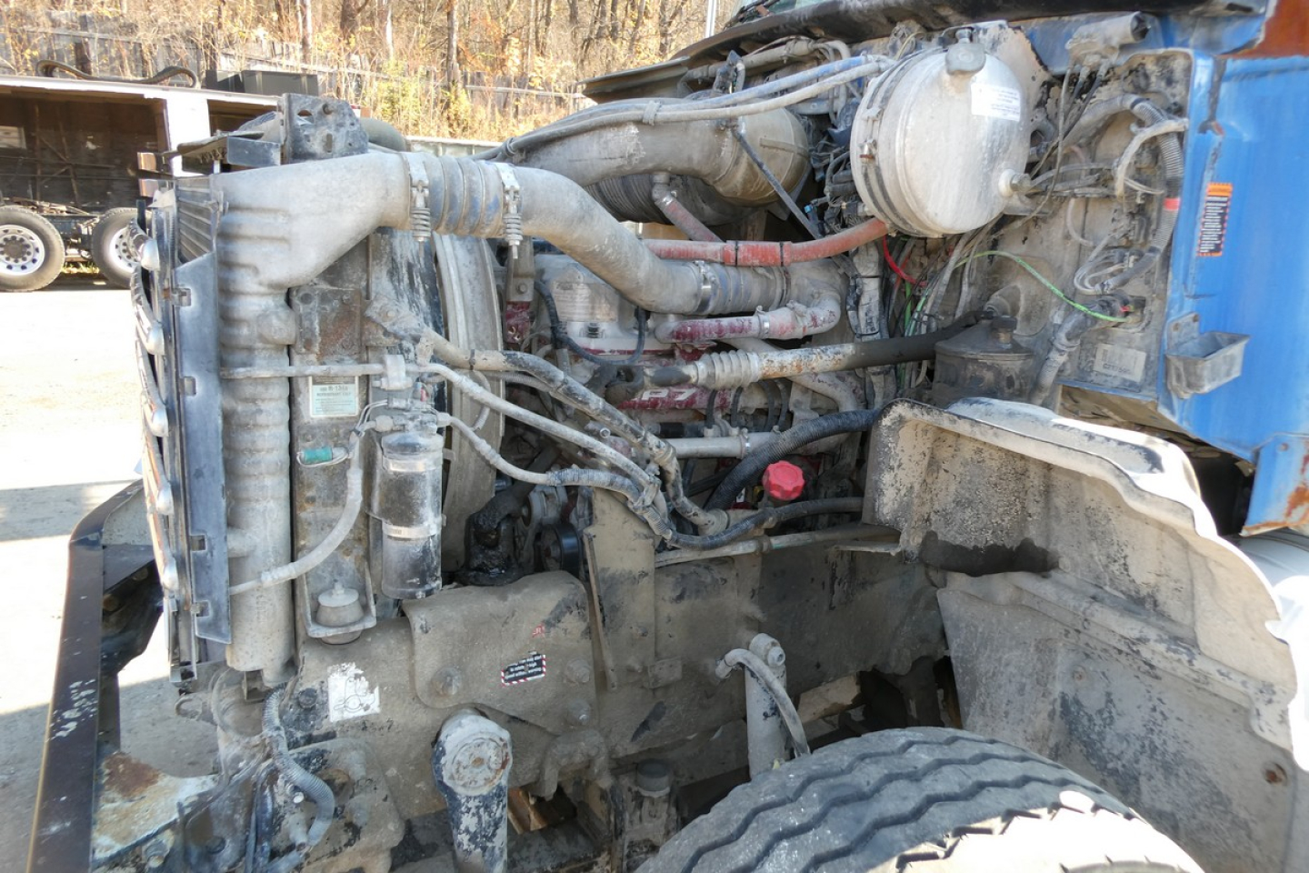 Mack truck engine bay with exposed components, hoses, and wiring in view from the left side; visible radiator, air intake, and various labels.