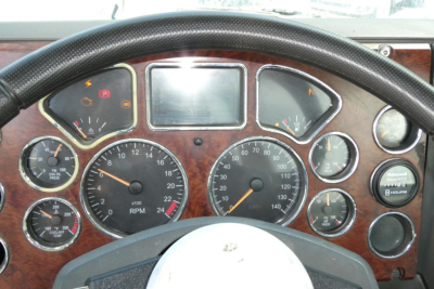 Mack truck cockpit/dashboard with multiple gauges and wooden panel; steering wheel visible; central digital display; various dials including RPM, speedometer, and warning lights.