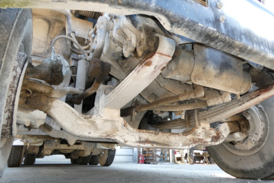 Underbelly view of a Mack mixer truck chassis showing suspension components, drive shaft, and frame members with dirt and grime.