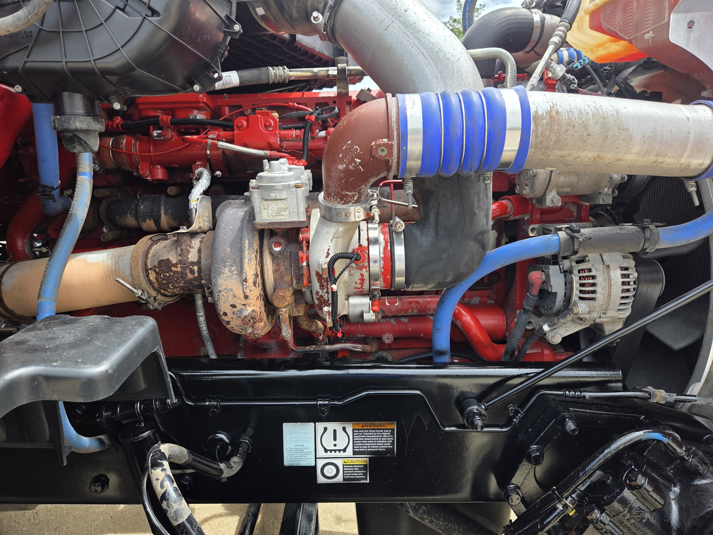 Peterbilt 567 engine bay showing red engine, blue coolant hose, turbocharger, and intake piping with metal clamps.