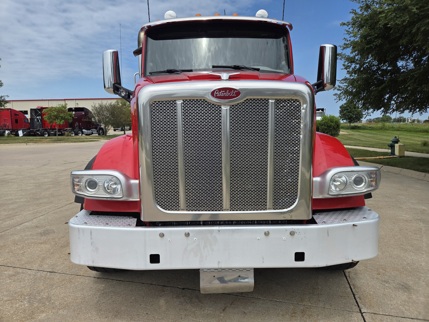 Peterbilt 567 front view showing large chrome grille, headlights, and bumper with emblem. Red cab, mirrors, and clear sky in background.
