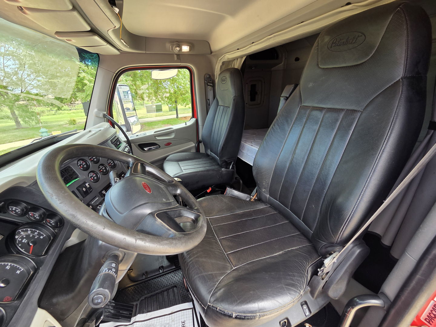Peterbilt 567 interior cab view with steering wheel, dashboard, and black leather seats; sleeper area visible in background.