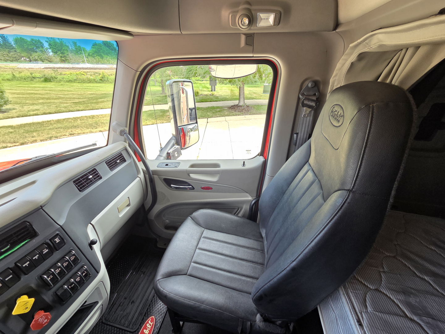 Truck interior front cab of a Peterbilt with steering wheel, driver's seat, and dashboard; visible text includes Peterbilt logo on seat, various switches, and a glimpse of exterior window.