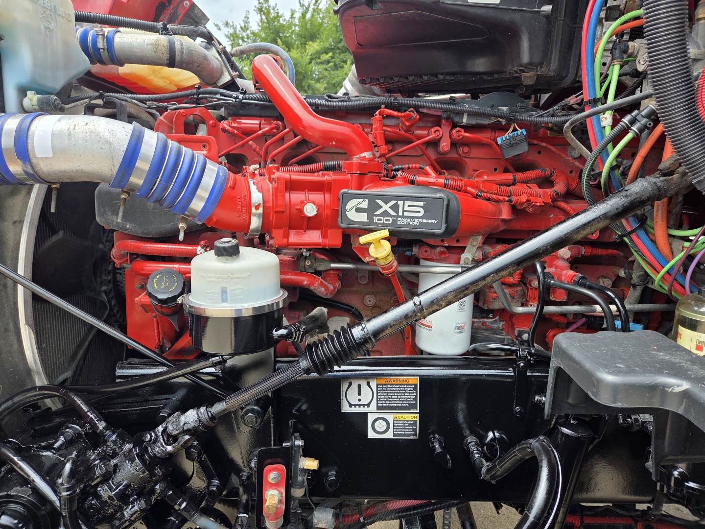 Peterbilt 567 engine bay showing Cummins X15 engine with red paint, various hoses, reservoirs, and wiring harnesses.