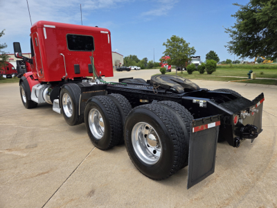 Peterbilt 567 conventional sleeper tractor with red cab, three rear axles, and fifth-wheel, viewed from the side in a parking lot.