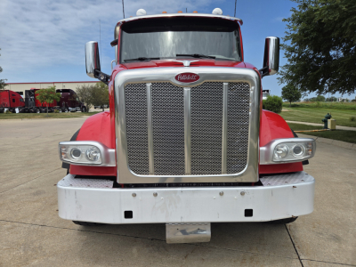 Peterbilt 567 front view showing large chrome grille, headlights, and bumper with emblem. Red cab, mirrors, and clear sky in background.