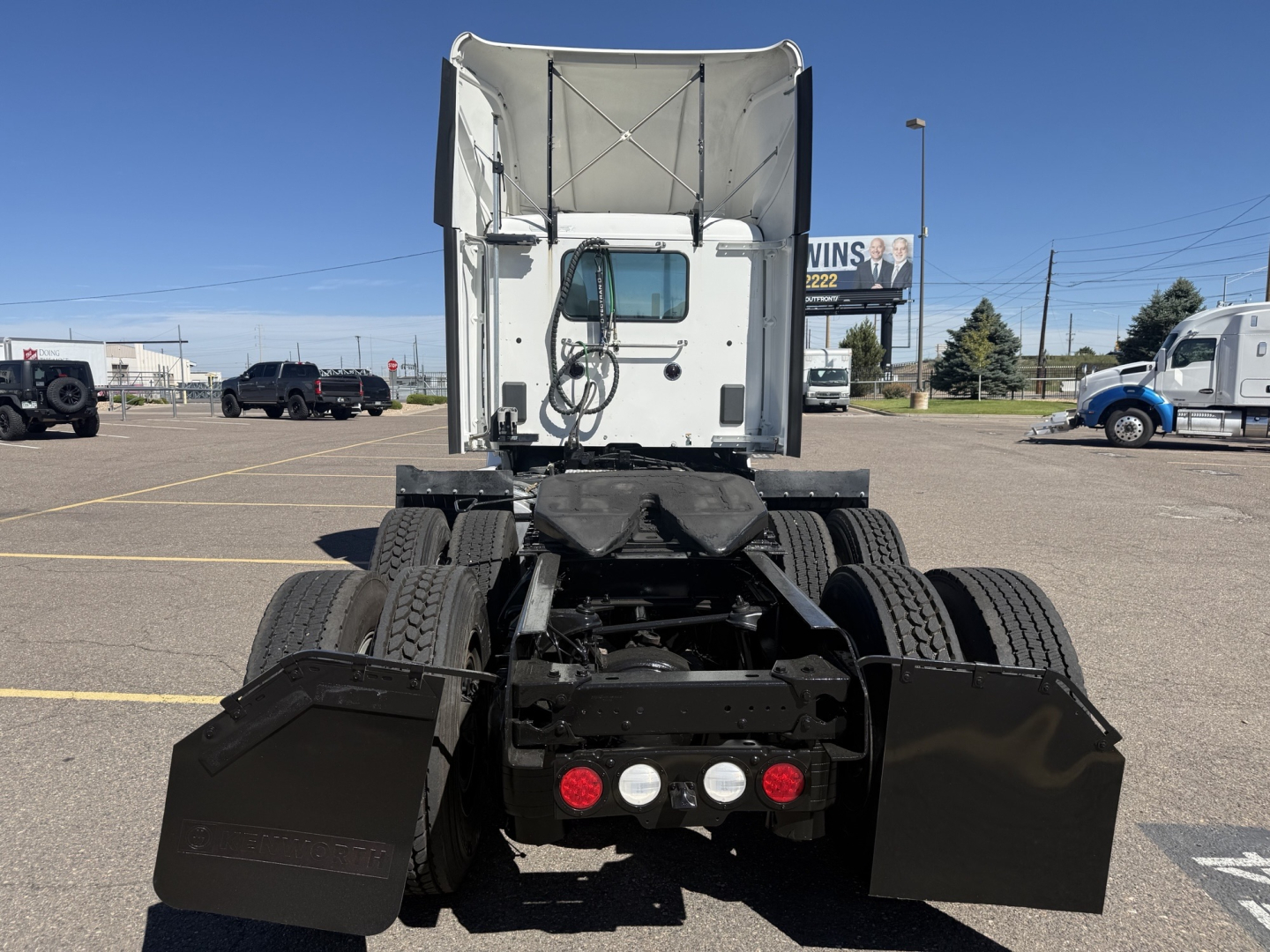 Kenworth T680 rear view showing fifth wheel, dual rear axles, mud flaps, and exhaust stack with white cab in background; no text visible on components.