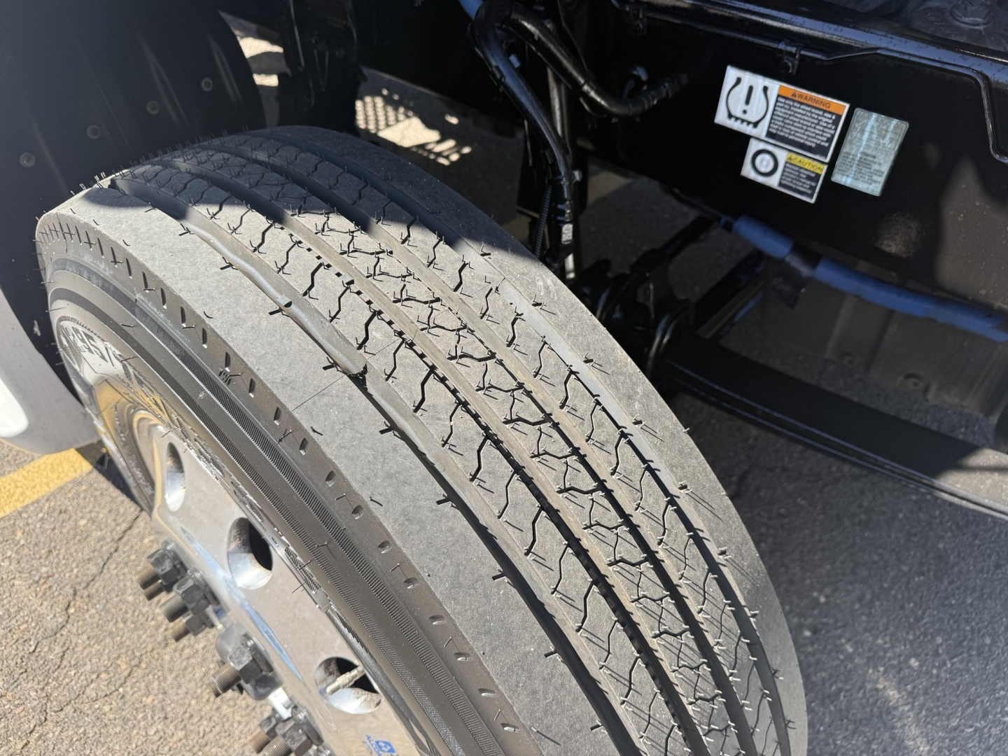 Tire and wheel on Kenworth truck chassis with warning labels and untermined hoses in the engine bay area.