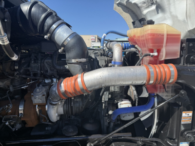 Kenworth T680 engine bay showing coolant reservoir with orange hose, air intake piping, and various hydraulic lines.