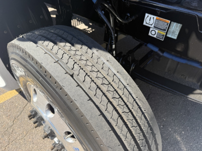 Tire and wheel on Kenworth truck chassis with warning labels and untermined hoses in the engine bay area.
