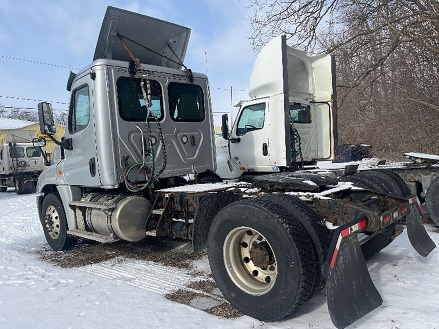 Truck cab and chassis of a Freightliner Cascadia125 tractor in a yard, snow on ground, two cabs visible, left side shows diesel fuel tank and steps, open hood area.