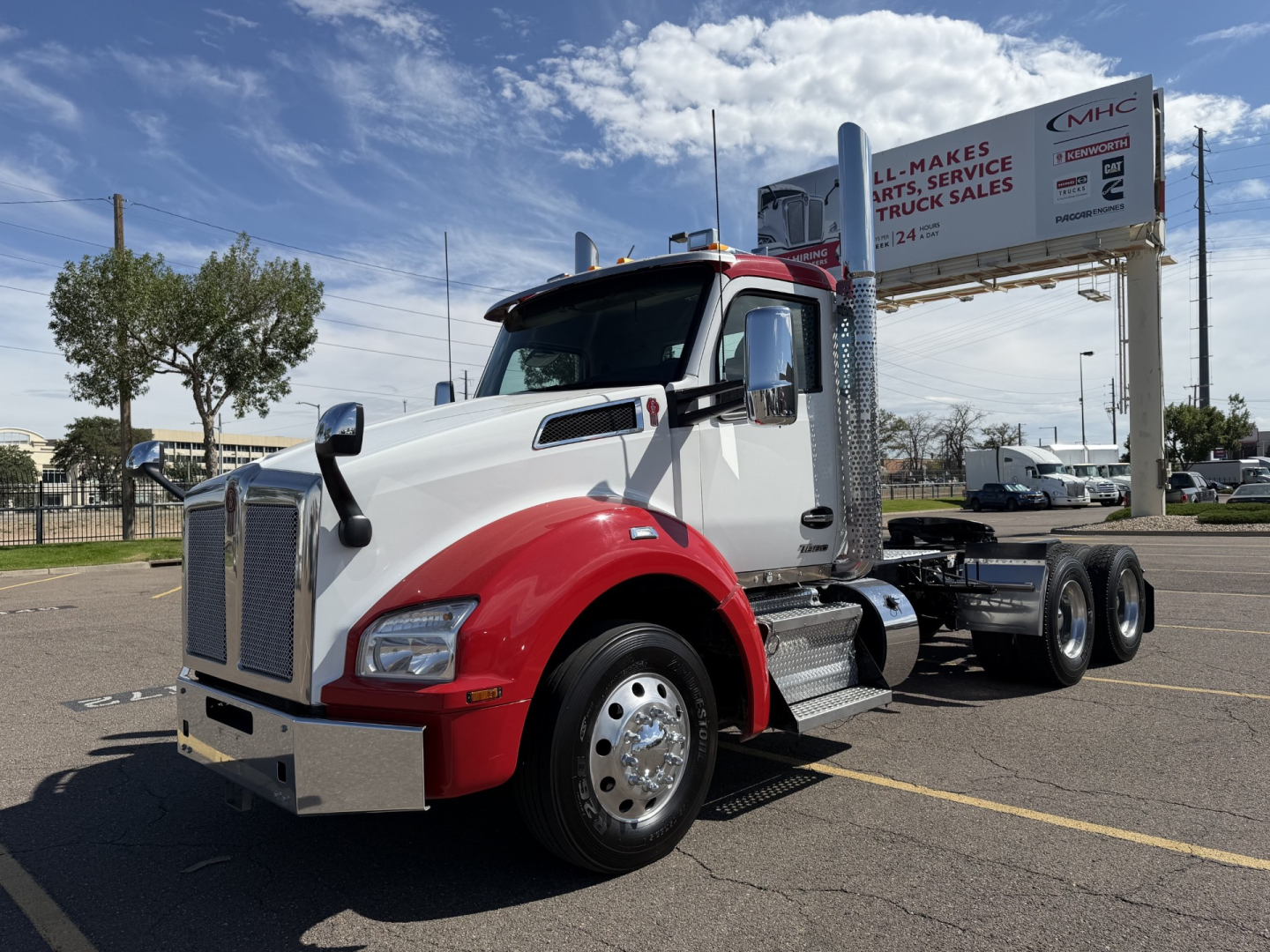 Kenworth T880 daycab tractor with white cab and red fenders, showing a large grille and dual rear wheel configuration; background includes signboard and parking lot.