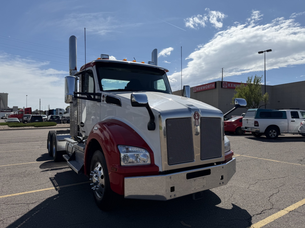 Kenworth T880 tractor in a parking lot, front view with chrome grille, two tall exhaust stacks, maroon lower fenders, and large chrome bumper.