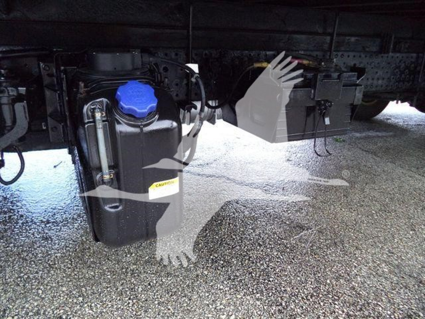 Underbody view of a 2015 Isuzu NPR HD chassis showing a blue engine coolant/def reservoir cap, hoses, and nearby metal brackets on a truck frame.