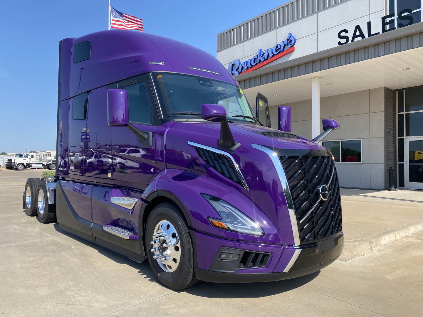 Truck in a purple Volvo VNL64T860 sleeper cab showing front grille and side mirrors in front of dealership building.