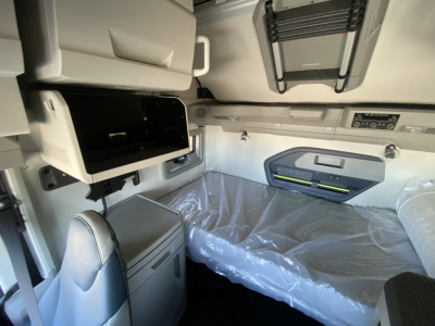 Volvo VNL cab interior showing sleeper bunk with plastic-covered bed, overhead storage, and control panel. Visible components include small open cabinet, fold-down table, and window shade.