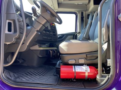 Cab interior of a new Volvo VNL64T860 tractor-trailer showing steering wheel, dashboard, driver seat, and a red fire extinguisher on the floor.