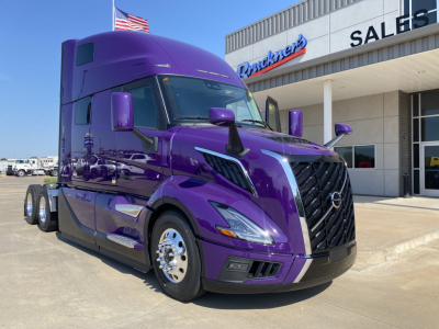 Truck in a purple Volvo VNL64T860 sleeper cab showing front grille and side mirrors in front of dealership building.
