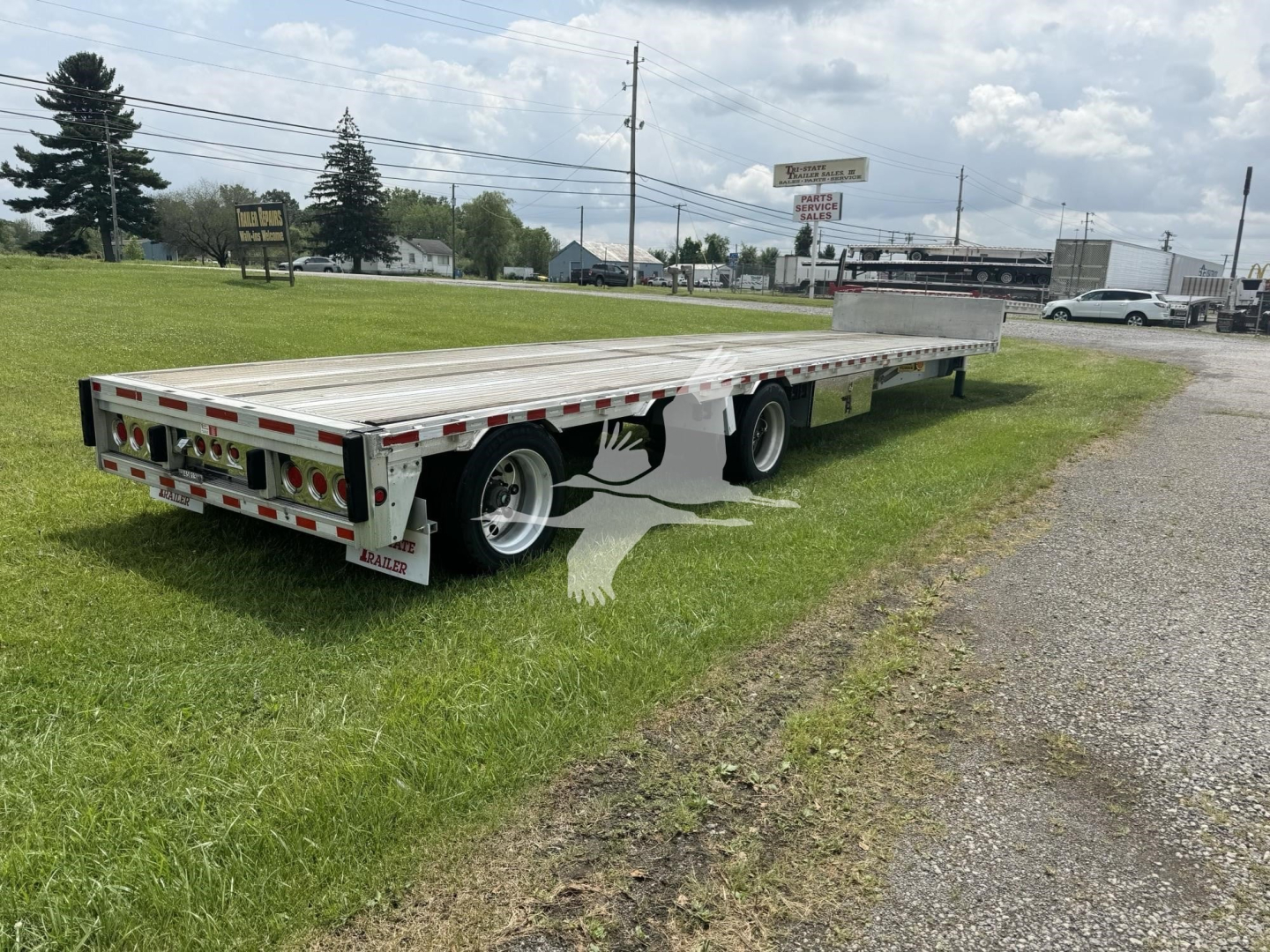 Flatbed trailer showing a long drop deck with aluminum surface and rear lighting, side rails, and triple-axle configuration.