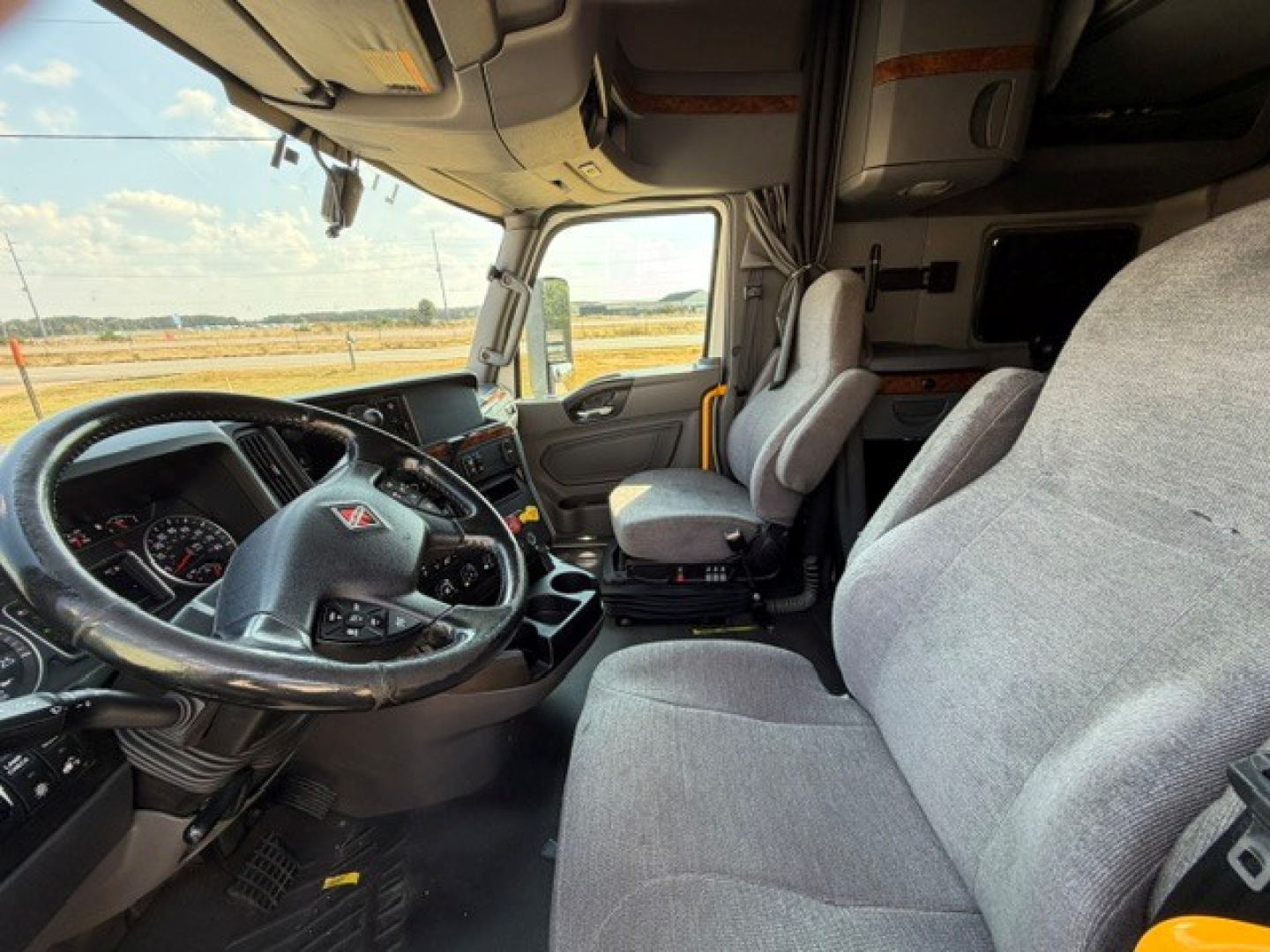 Truck cab interior showing steering wheel with dashboard, driver seat, passenger seat, and center console. Grays fabric seats, steering wheel with various controls, windows, and a glimpse of exterior highway.
