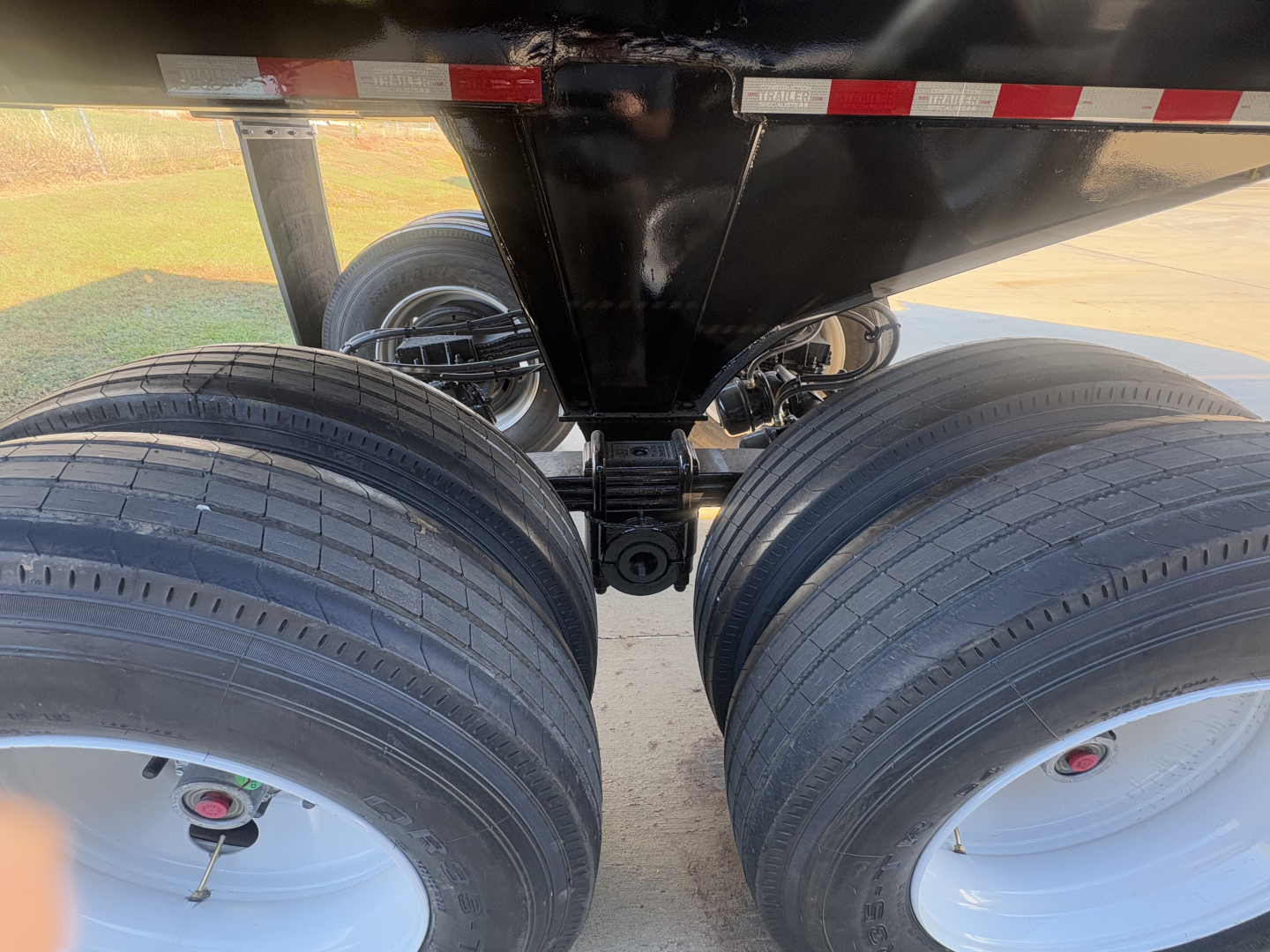 Trailer axles and dual-tire configuration under a dump trailer showing two axles with black tires and white rims, connected by a hinge and undercarriage frame.