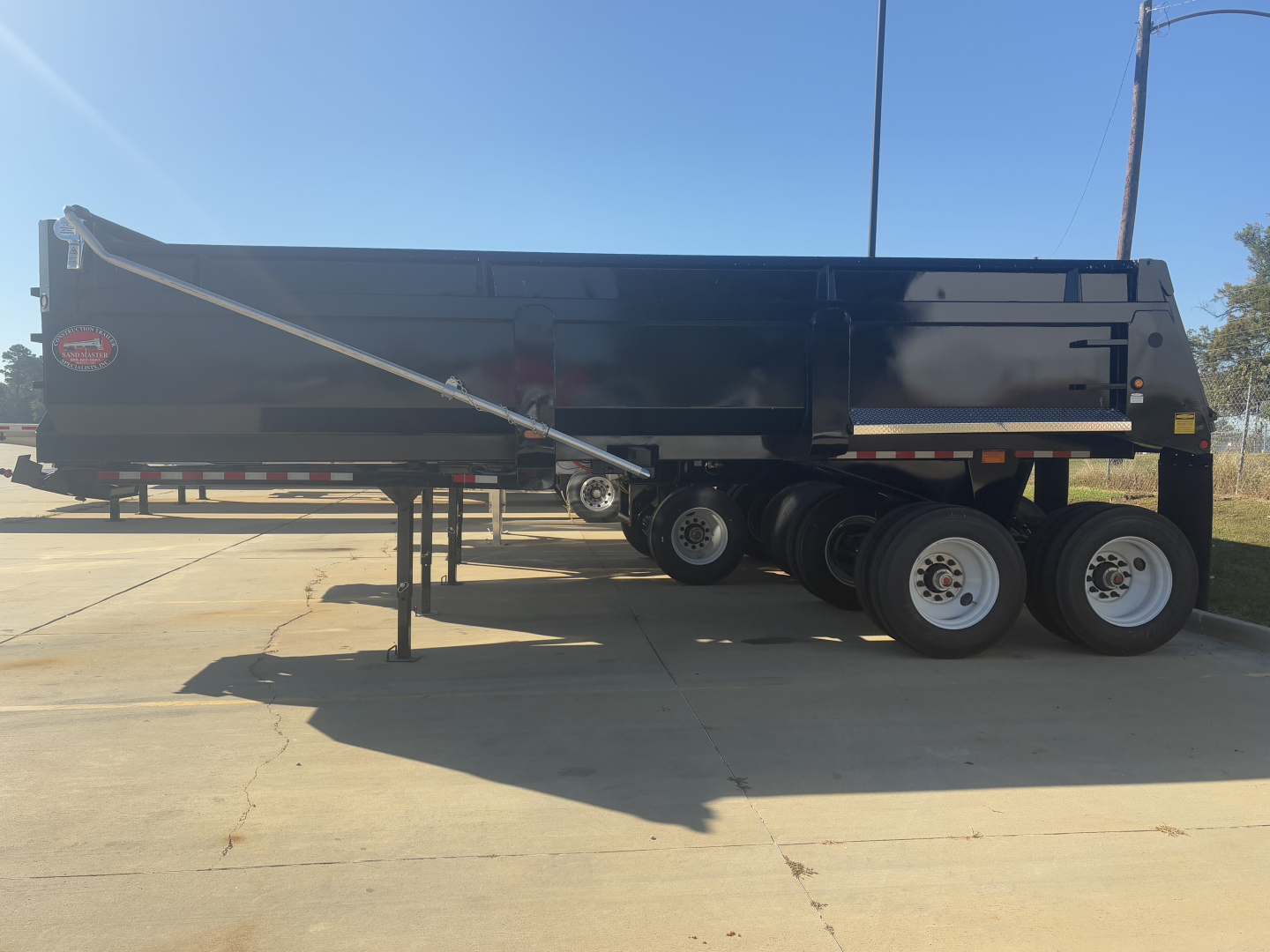 Dump trailer side view with black steel body and triple axle, wheel mudflaps, and support legs extended.