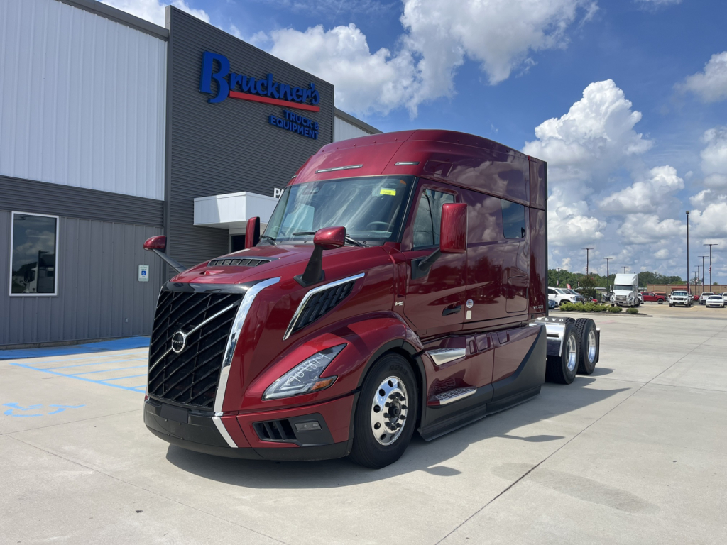 Volvo VNL64T840 tractor-trailer in front of dealership, red exterior, Sleeper cab, dual rear axles, close-up of front grille and headlights, dealership sign in background.