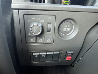 Dashboard controls of a Volvo VNL64T840 showing headlight switch, engine start/stop button, and several switches; portion of instrument cluster reflected in the window.