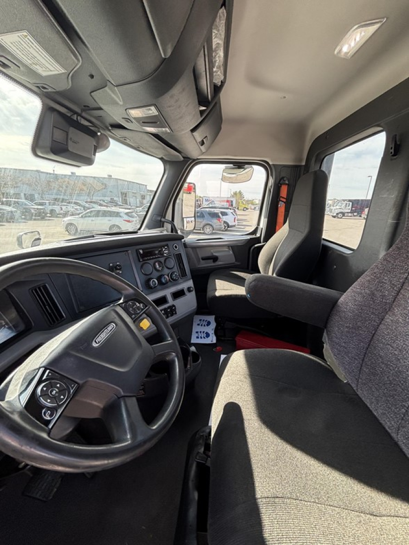 Cab interior of a Freightliner Cascadia showing steering wheel, dashboard with gauges, driver's seat, and passenger seat. Visible controls on the steering wheel and center console.