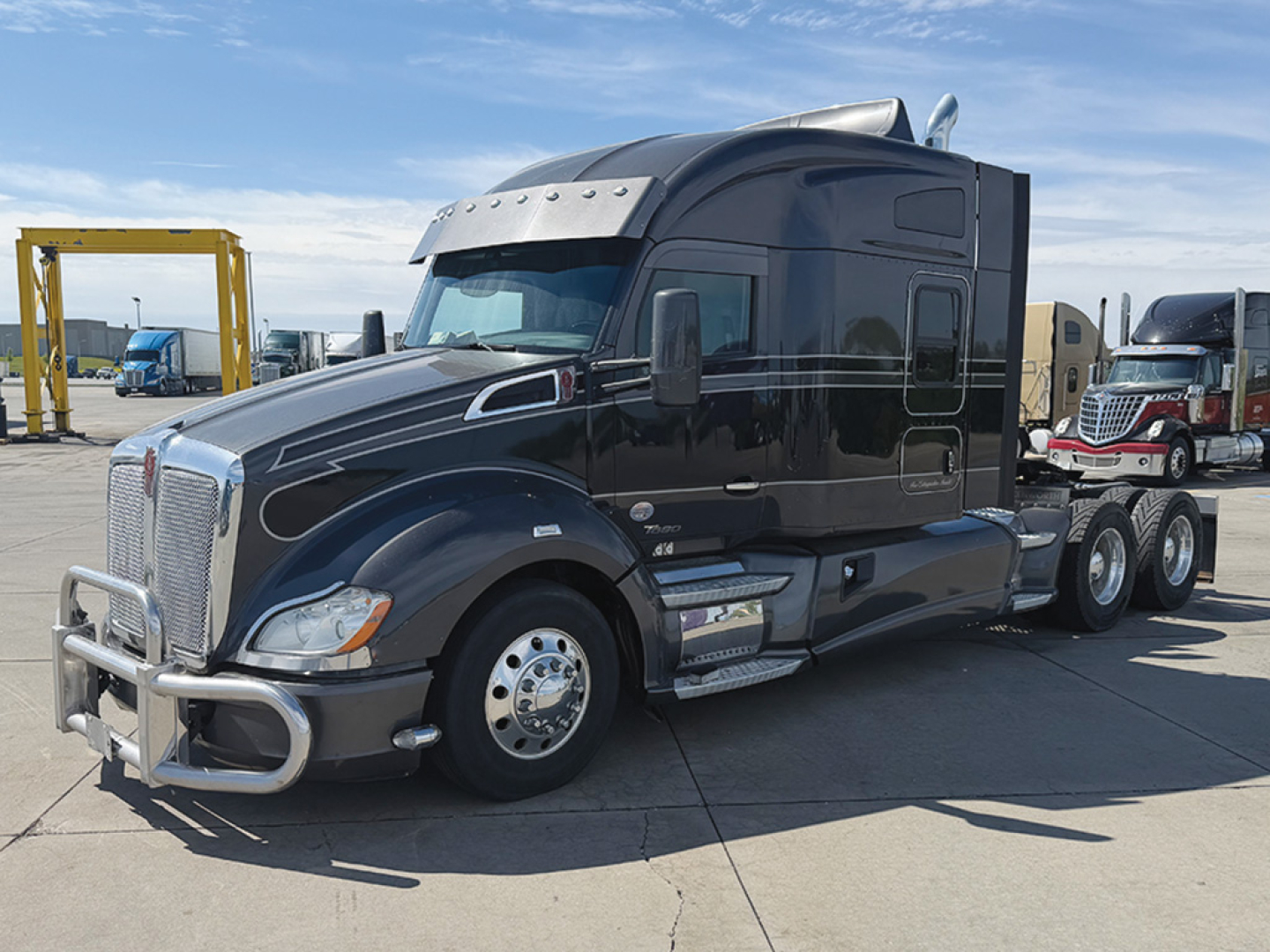 Kenworth T680 conventional sleeper truck in dark color with chrome grille guard and sun visor, side exhaust, dual rear axles, in a dealership lot.