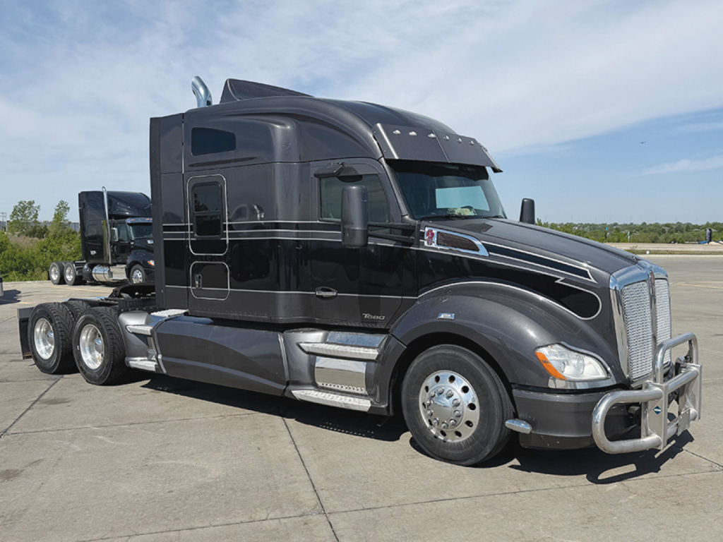 Kenworth T680 sleeper cab tractor in dark color, side view, three rear axles, chrome bumper guard, large exhaust stacks, highway tires.