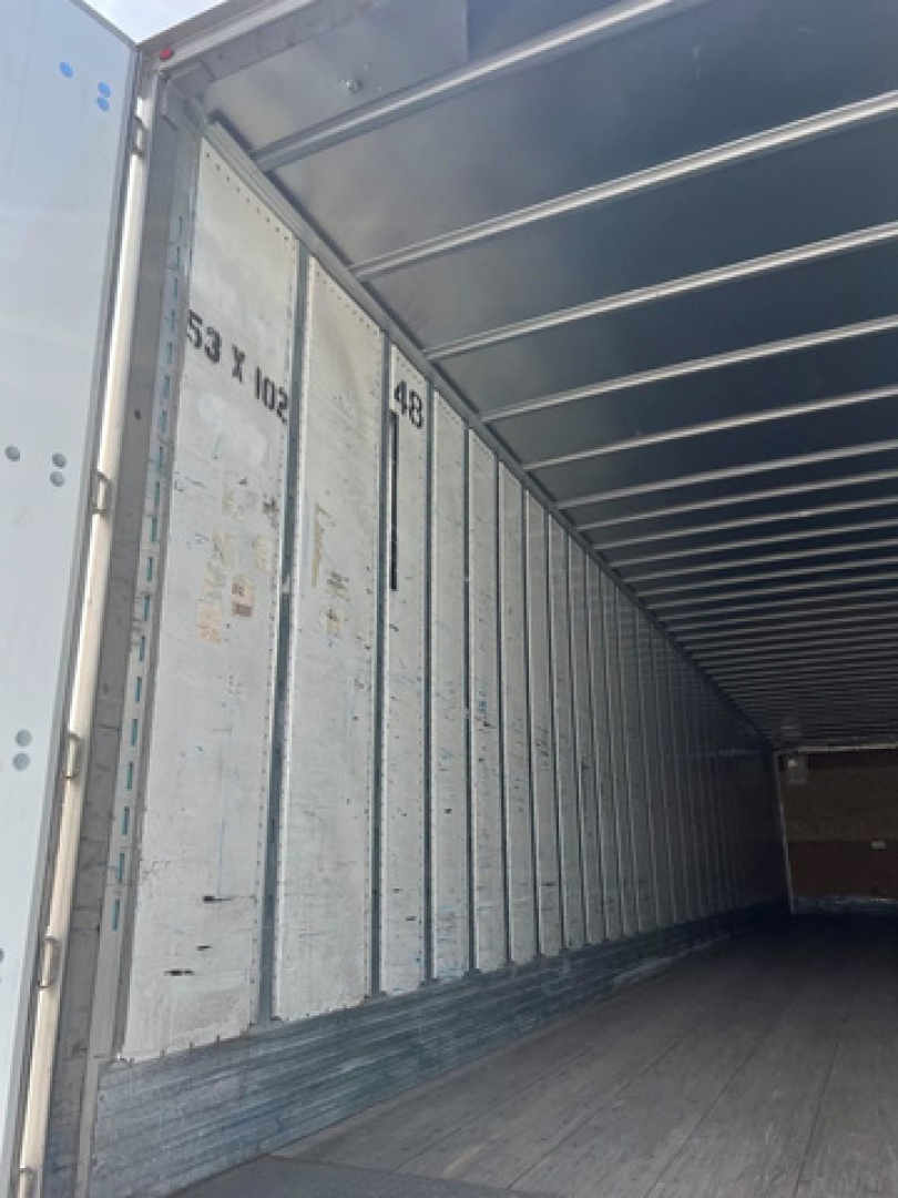 Interior of a utility van trailer showing metal side panels with vertical ribs and a wooden floor. Numbers 53 x 102 appear on a panel. Open rear door framing visible.