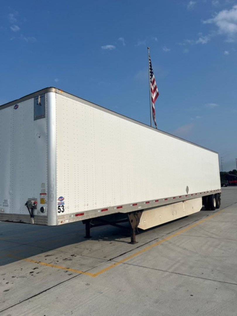 Long white utility trailer with a raised landing gear and two axles, parked on a concrete lot with a flagpole and American flag in the background.