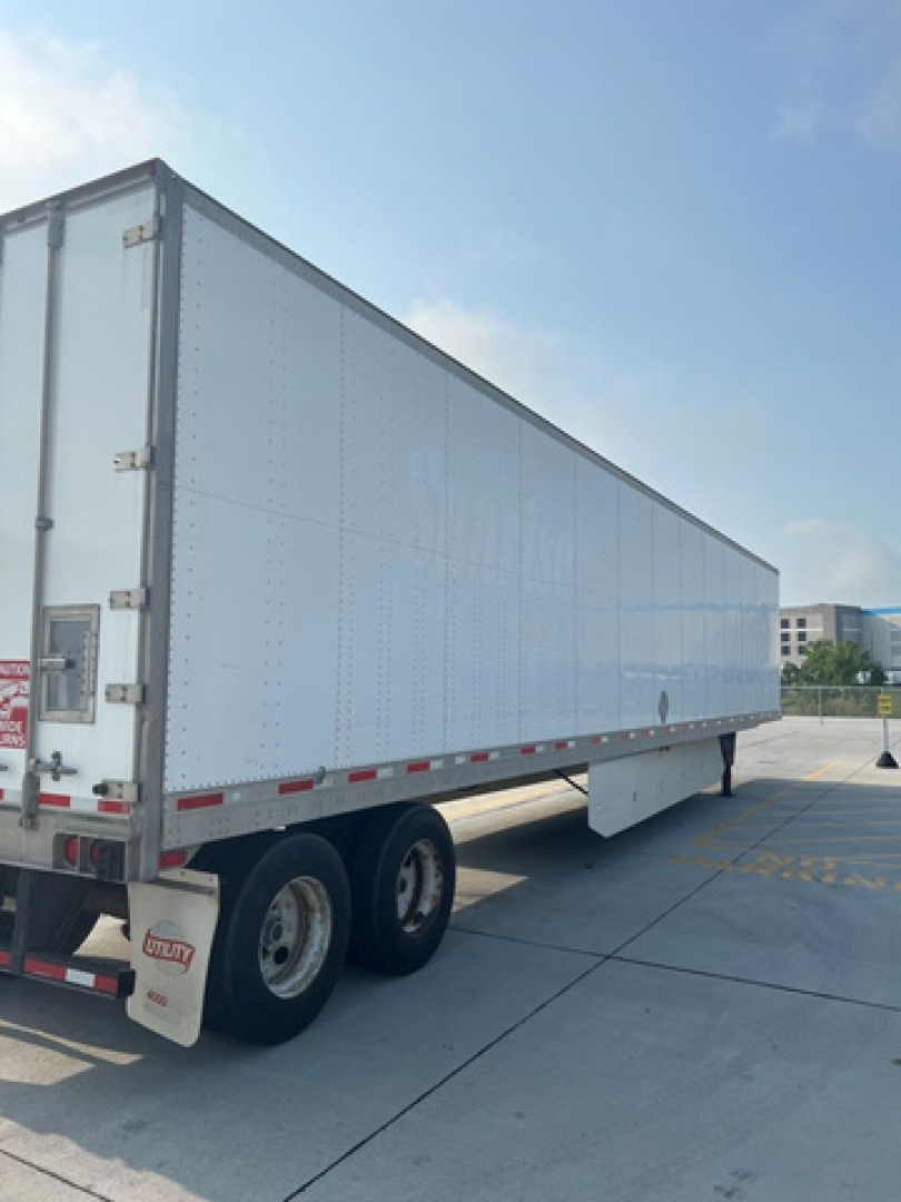 Utility van trailer in a paved lot showing a large white box trailer with tandem rear wheels.