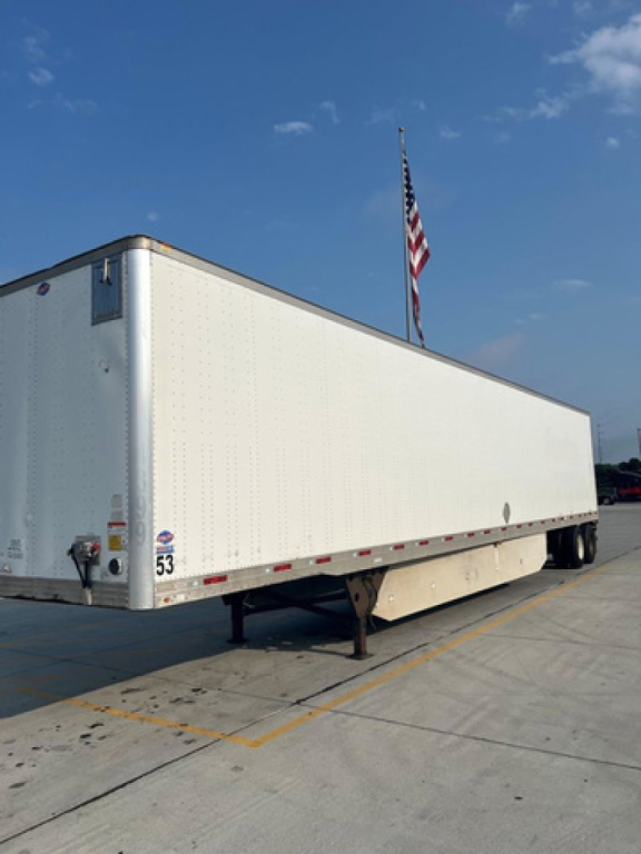 Long white utility trailer with a raised landing gear and two axles, parked on a concrete lot with a flagpole and American flag in the background.