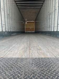 Trailer interior showing empty cargo space with wooden floor and metal sidewalls; front wall with wooden bulkhead in distance.