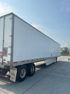 Utility van trailer in a paved lot showing a large white box trailer with tandem rear wheels.